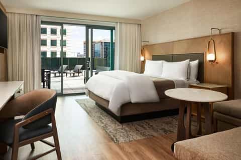 Modern hotel bedroom with king bed, wood headboard, balcony access, and city views
