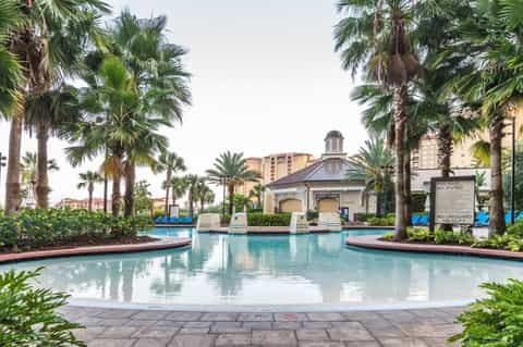 Luxurious resort pool surrounded by palm trees with colonial-style pavilion and high-rise buildings in background