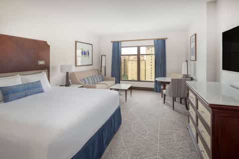 Spacious hotel room with king bed, seating area, and balcony overlooking city buildings