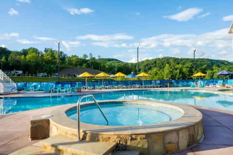 Outdoor swimming pool with hot tub, yellow umbrellas, and forested hills in the background on sunny day