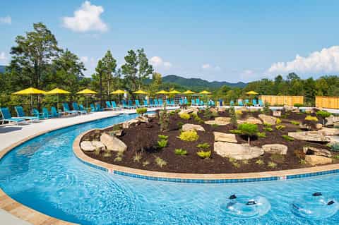 Curved resort pool with blue loungers, yellow umbrellas, and landscaped island featuring rocks and plants with mountain views