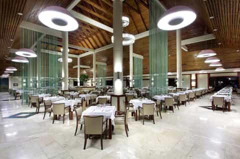 Spacious modern dining hall with circular pendant lights, wooden ceiling beams, white tablecloths, and beige upholstered chairs