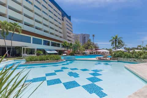 Resort pool with blue and white checkered tile pattern, palm trees, and multi-story hotel building under clear sky