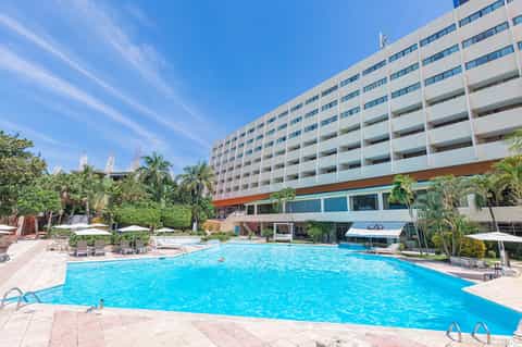 Large outdoor swimming pool with bright blue water, surrounded by lounge chairs and tropical palm trees, with multi-story hotel building in background