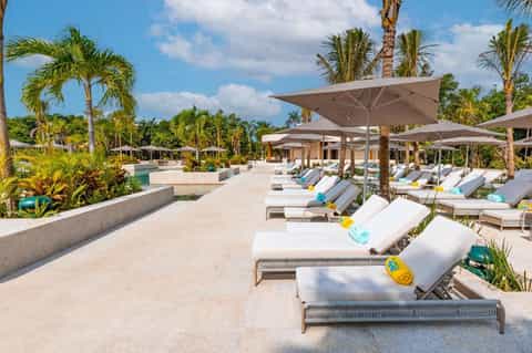 Luxury resort pool deck with white lounge chairs, large umbrellas, and palm trees under blue sky