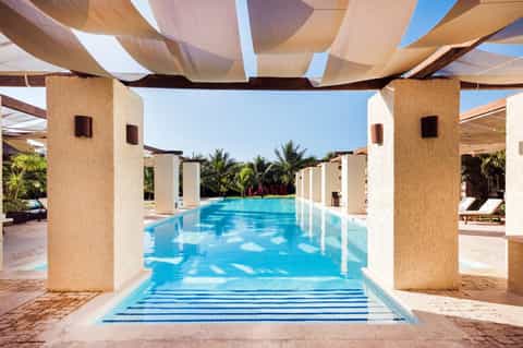 Covered walkway with white columns leading to bright blue swimming pool surrounded by palm trees