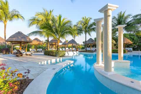 Luxury resort pool with thatched-roof cabanas, stone columns, and palm trees under clear sky