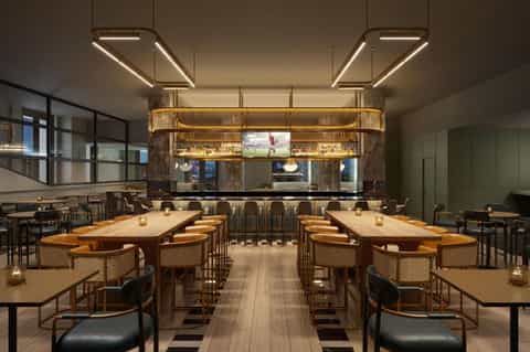 Modern hotel lounge with wooden tables, tan chairs, geometric lighting fixtures, and a gold-framed bar with TV display