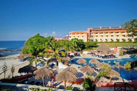 Beachfront resort with orange and white buildings, pool with tiki umbrellas, palm trees, and ocean view