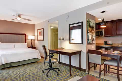 Modern hotel suite with queen bed, work desk, kitchenette, and striped area rug in warm tones