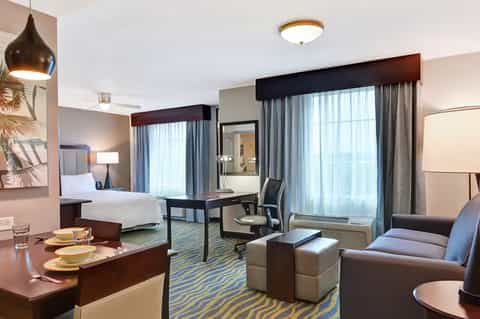 Modern hotel suite with bed, work desk, dining area, and floor-to-ceiling windows with city views