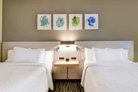 Modern hotel room with two queen beds, white linens, neutral headboard, and four colorful abstract watercolor paintings above