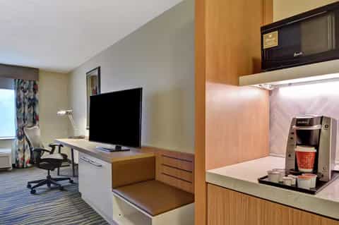 Modern hotel room with work desk, flat-screen TV, coffee maker, and wood cabinetry in neutral tones