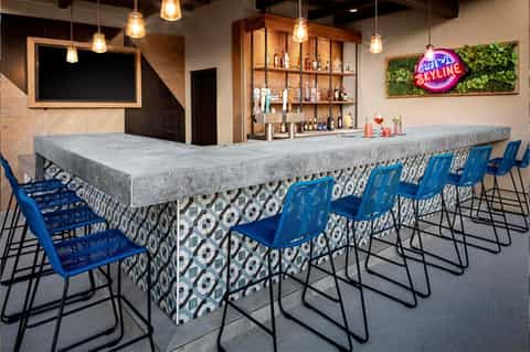 Modern bar with concrete countertop, patterned base, blue chairs, pendant lights, and neon sign