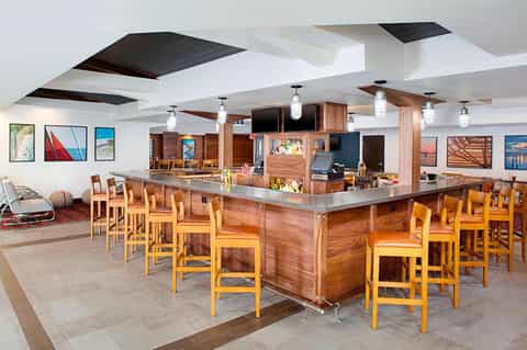 Spacious hotel bar and lounge with wooden counter, bar stools, pendant lighting, and artwork on white walls