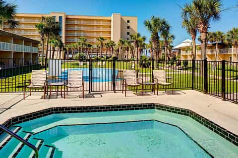 Outdoor heated pool with lounge chairs surrounded by palm trees and multi-story beige resort buildings