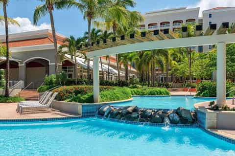 Resort pool with waterfall feature, pergola, tropical landscaping, and contemporary architecture in daylight