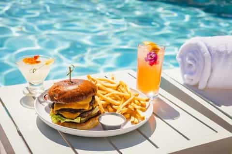 Gourmet burger with fries and cocktails poolside on white deck next to towel