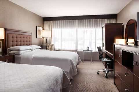 Hotel room with two queen beds, brown headboards, desk area, and floor-to-ceiling windows with sheer curtains