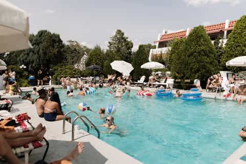 Outdoor resort pool filled with guests, white umbrellas, and Spanish-style buildings in background