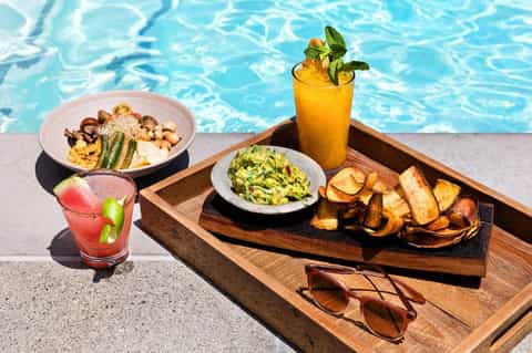 Poolside dining setup with colorful cocktail, seafood pasta, guacamole, and roasted potatoes on wooden trays