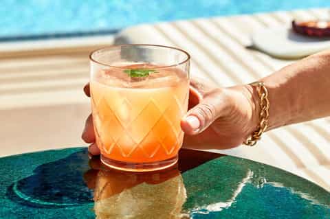 Guest holding coral-colored cocktail with mint garnish by poolside