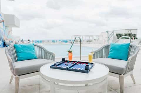 Two white modern chairs with turquoise pillows facing round table with backgammon board on beach terrace
