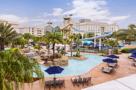 Resort water park with lazy river, slides, palm trees, and multi-story hotel buildings under blue sky
