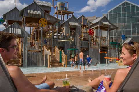 Couple relaxing poolside at water park with rustic wooden play structures and splash pad