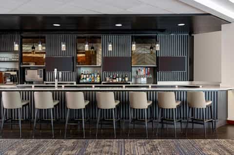 Contemporary hotel bar with black corrugated metal backdrop, pendant lighting, and cream-colored bar stools