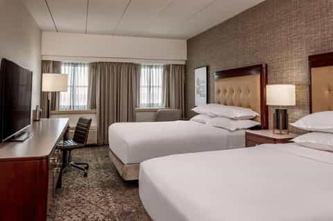Hotel room with two queen beds, wood furniture, textured tan walls, and windows with beige curtains