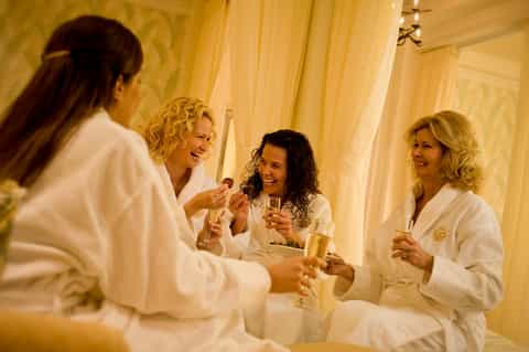 Four women in white robes toasting with champagne in a luxurious spa lounge with golden curtains