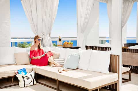 Woman in red dress relaxing on white cushioned lounge under sheer curtains with ocean view