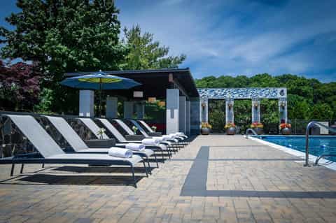 Resort pool deck with striped lounge chairs, blue umbrella, modern pavilion, and manicured gardens