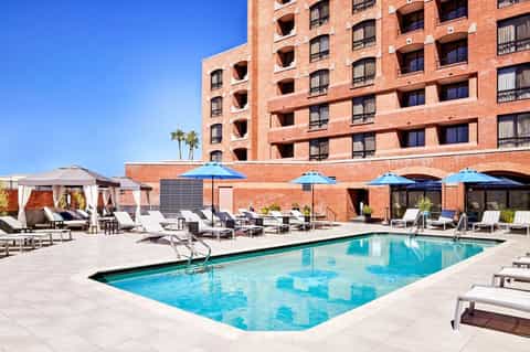 Brick hotel building with blue umbrellas and lounge chairs surrounding rectangular swimming pool under clear sky