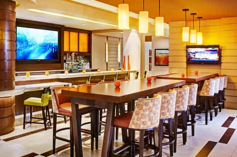 Upscale lounge with orange walls, bar seating, dining table, pendant lights, and multiple TV screens