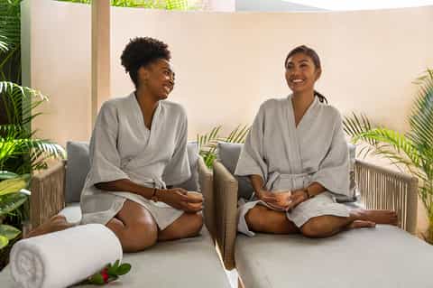 Two women in spa robes sitting on massage beds in a plant-filled wellness room