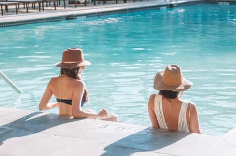 Two women in sun hats relaxing by a clear blue swimming pool on a sunny day