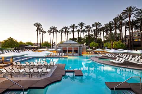 Large resort pool surrounded by palm trees, lounge chairs, and umbrellas with poolside cabana buildings