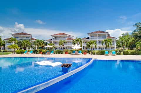 Luxury beachfront resort with multi-story white buildings, palm trees, and large blue swimming pool under clear sky