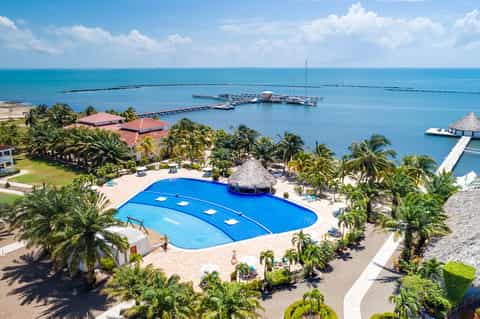 Aerial view of beachfront resort with blue pool, palm trees, and ocean pier under sunny sky