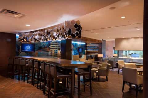 Hotel lounge with bar seating, geometric ceiling installation, wood flooring, and modern furnishings with blue accents