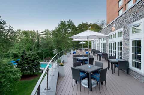 Modern deck with dining tables, umbrellas, and lounge seating overlooking a pool and lush green landscape