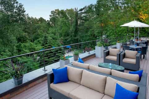 Elevated deck with rattan furniture, beige cushions, blue pillows, and forest views beyond metal railings