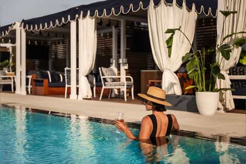 Woman in pool wearing straw hat, holding drink near white cabana with draped curtains