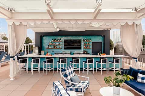 Rooftop bar with turquoise backdrop, white bar stools, cream curtains, and city skyline views