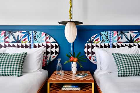 Modern hotel bedroom with twin beds, colorful patterned headboards, green gingham pillows, and pendant lighting