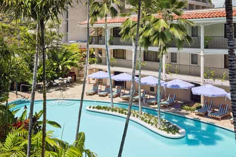 Resort courtyard pool surrounded by palm trees with white umbrellas and lounge chairs beneath multi-story hotel buildings