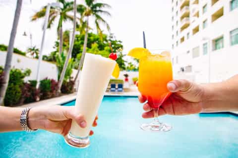 Two tropical cocktails held by pool with palm trees and resort buildings in background
