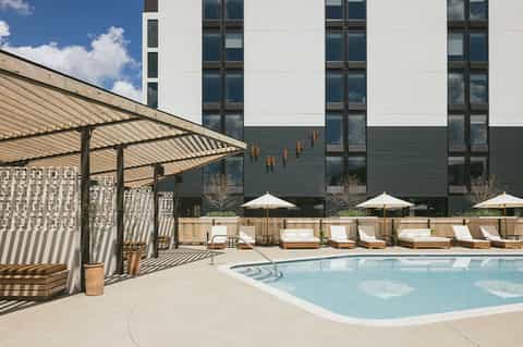 Modern hotel pool deck with loungers, umbrellas, pergola shade structure, and contemporary building facade
